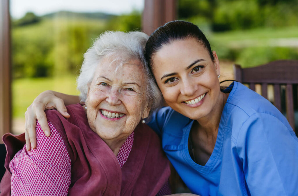 A caregiver and an older adult touch their faces together and smile at the camera while sitting on a porch together in senior living