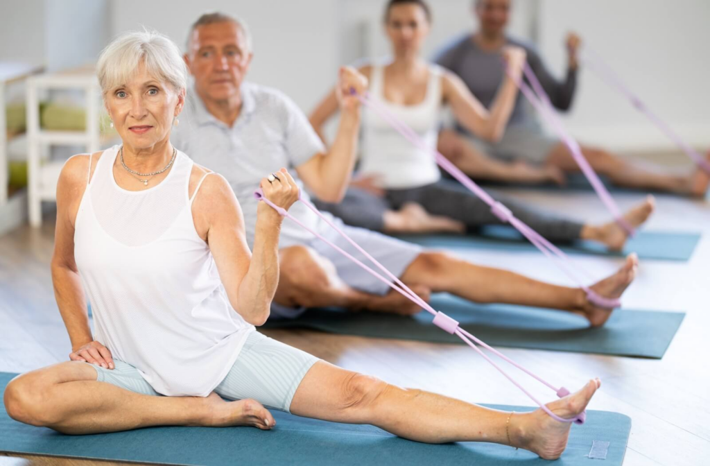 A group of older adults sit on the ground and use resistance bands around the edge of their foot to strengthen their legs