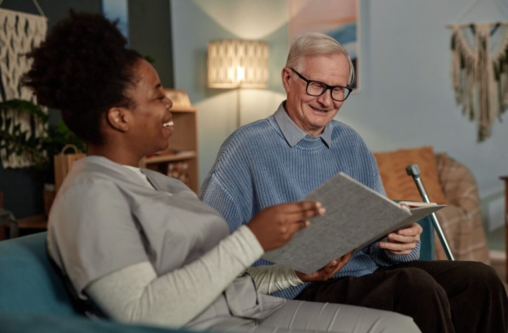 An older adult laughs while showing a smiling caregiver a family photo album while sitting together in assisted living.