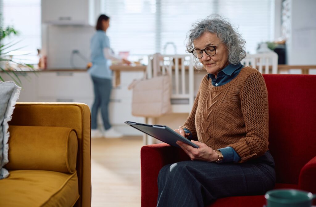 An older adult looks at a folder while sitting on a red armchair in their home in assisted living. In the background, a caregiver tidies their kitchen counters for them.