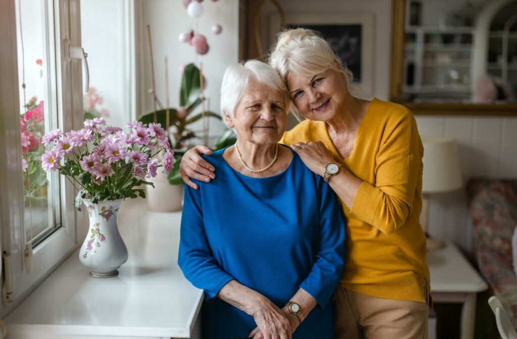 an older adult and their senior parent embrace and smile towards the camera in a cozy living room