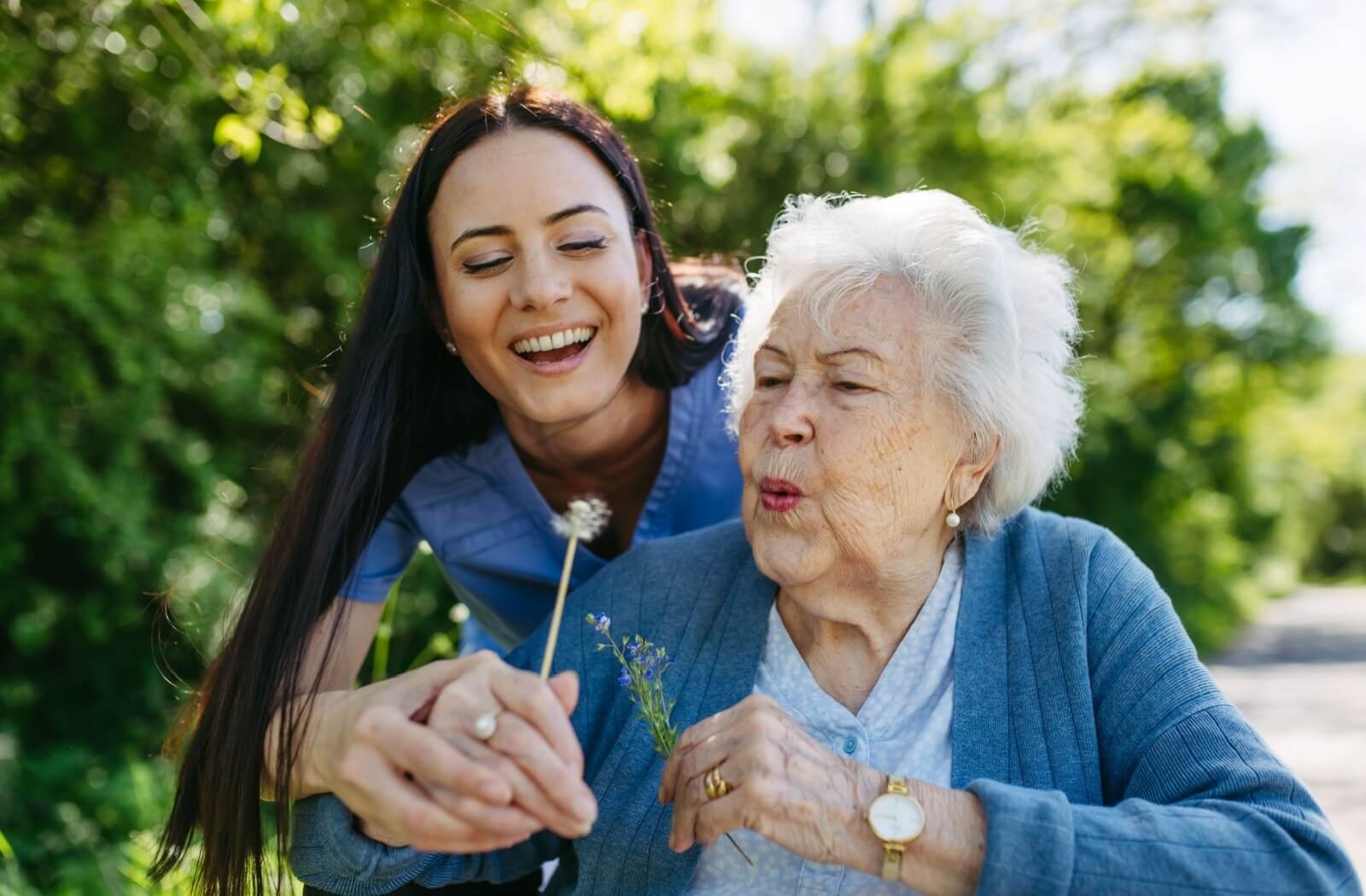 A senior blows the seeds off a dandelion with the help of a caregiver